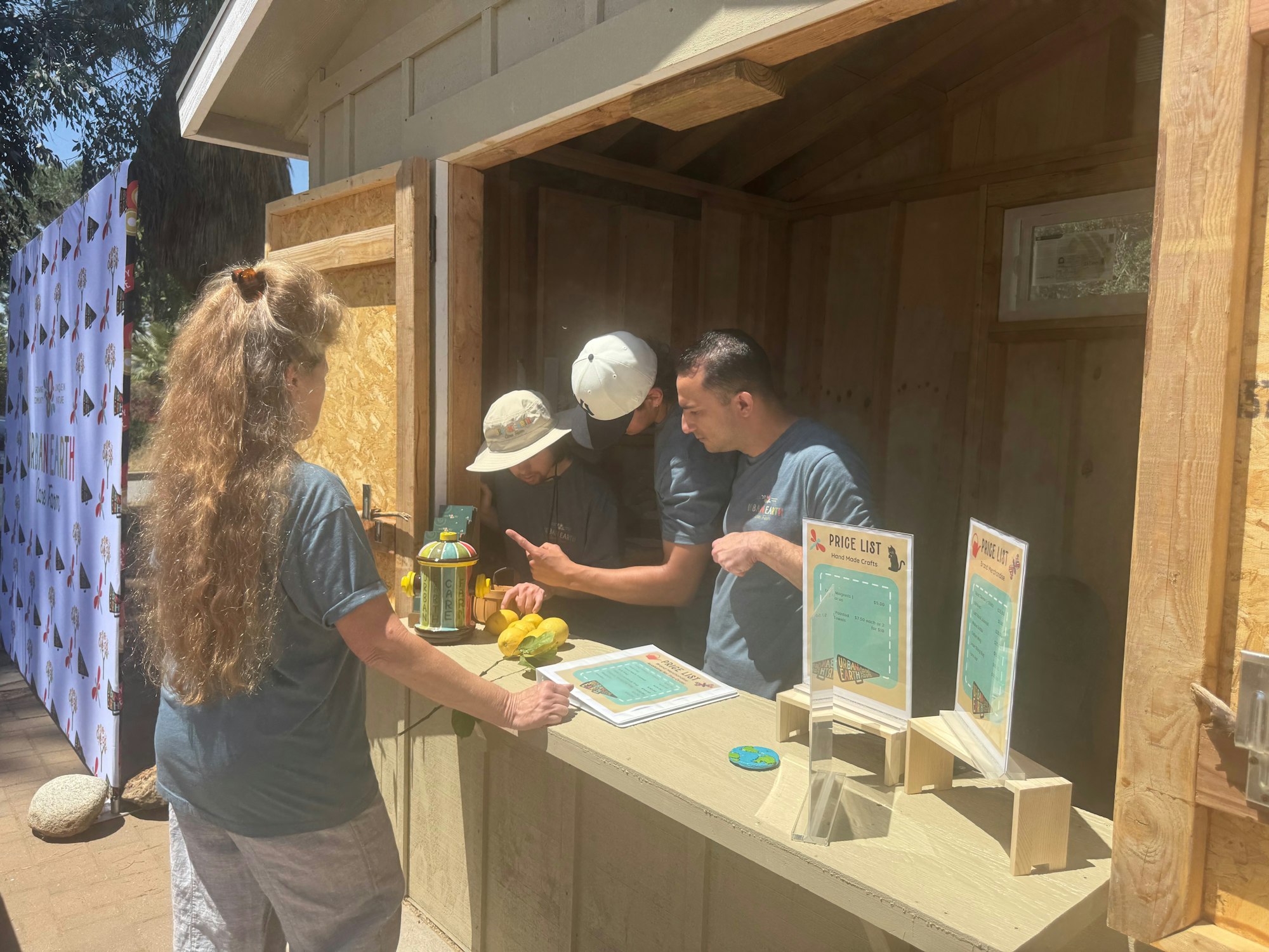 Three people are at a small wooden stand with a price list and lemons. One person with long hair is facing the booth.