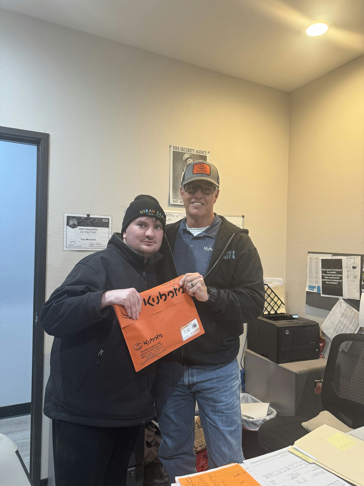 Two people in jackets and hats pose in an office holding an orange envelope.