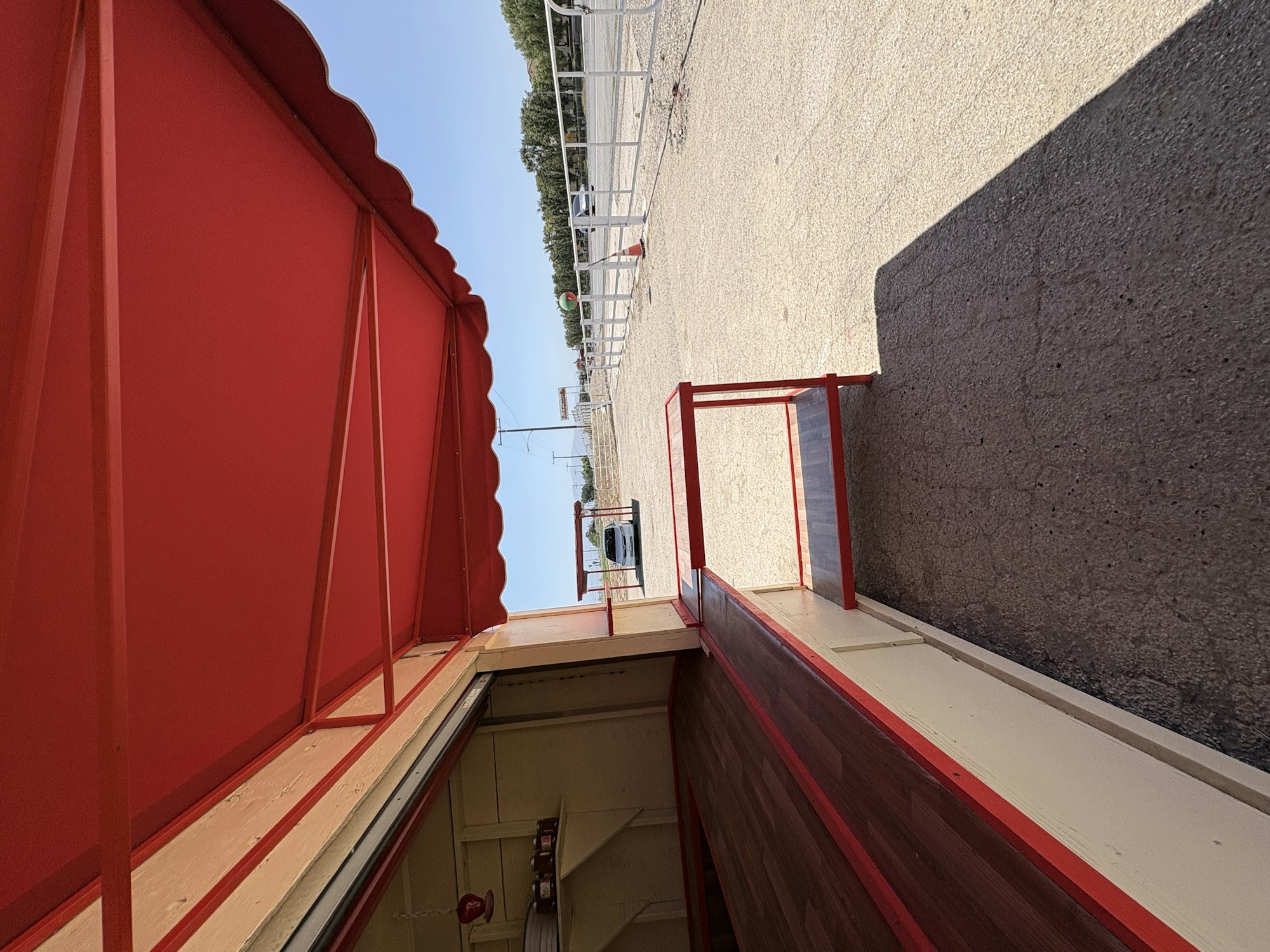 A red awning covers an open area, leading to a paved path with a glimpse of a landscape in the background.