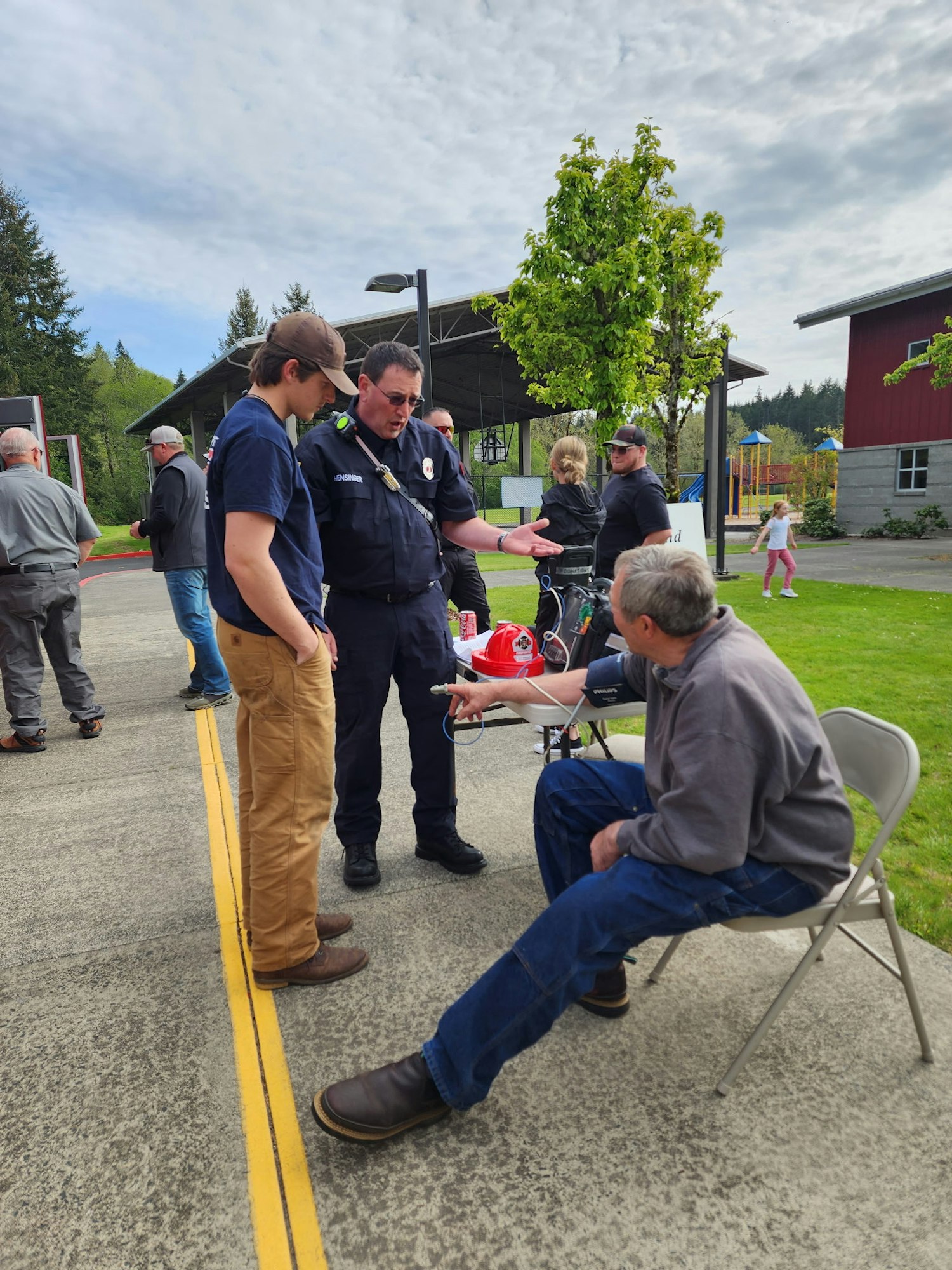 A group of people gathered outdoors, including a firefighter talking to two men, one seated, near a playground and building.