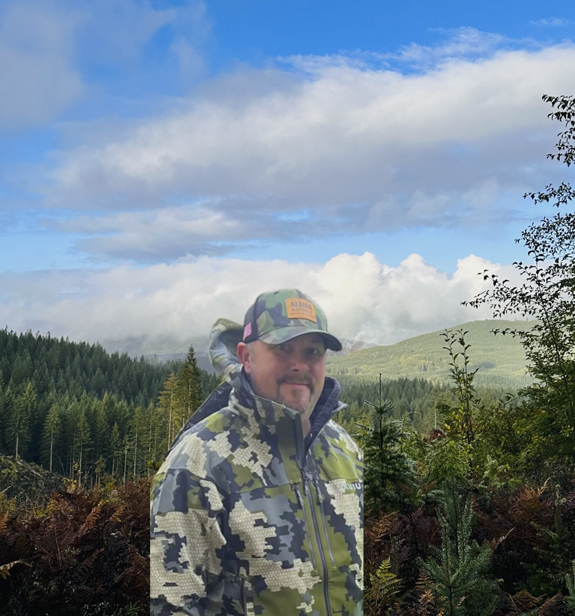 Man in camo jacket and cap with forest and clouds in the background.