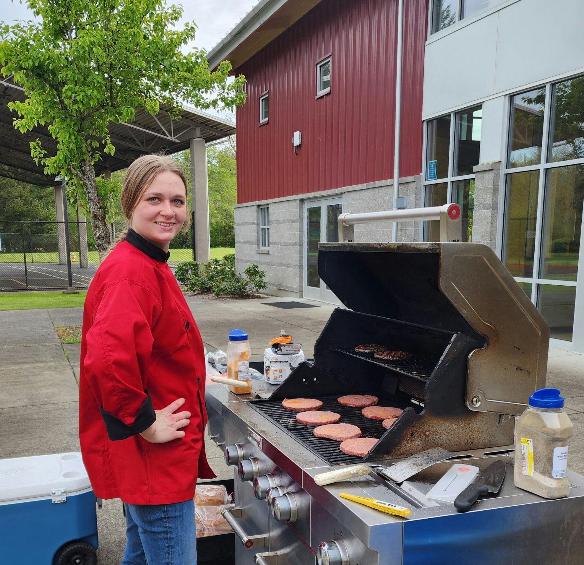 A person in a red chef coat is grilling burgers outdoors beside a building with a red wall.