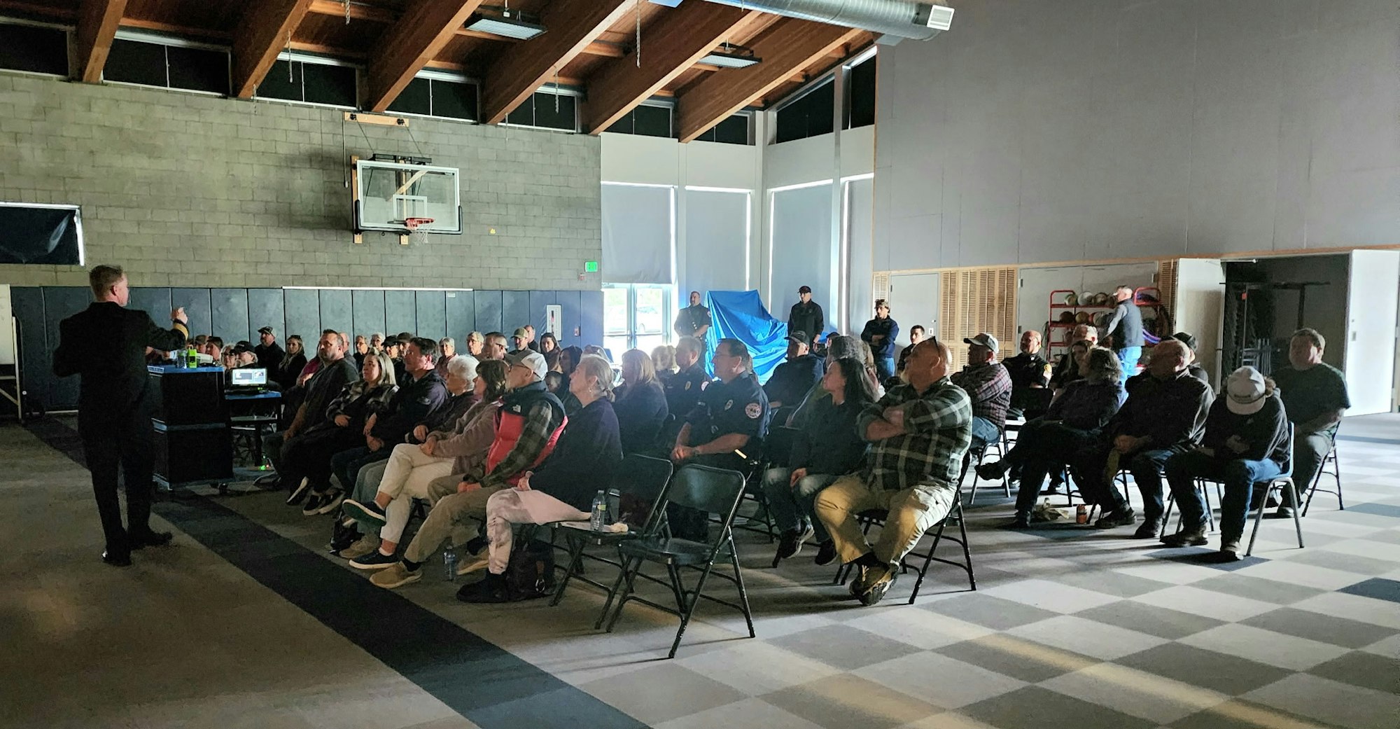 A speaker addresses an audience seated in a large room with a high ceiling and a basketball hoop.