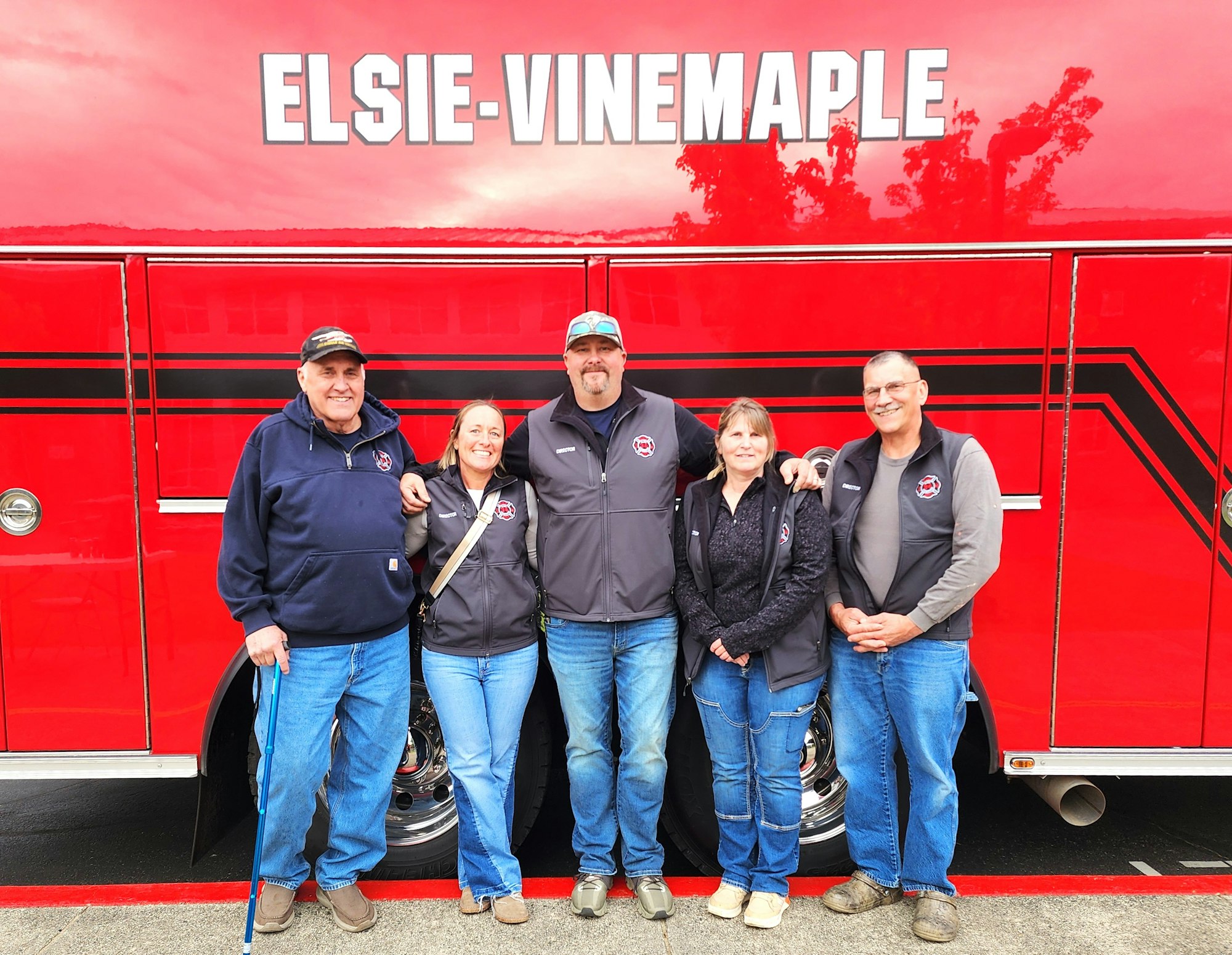 Five people in front of a red vehicle with "Elsie-Vinemaple" written on it, wearing matching jackets.