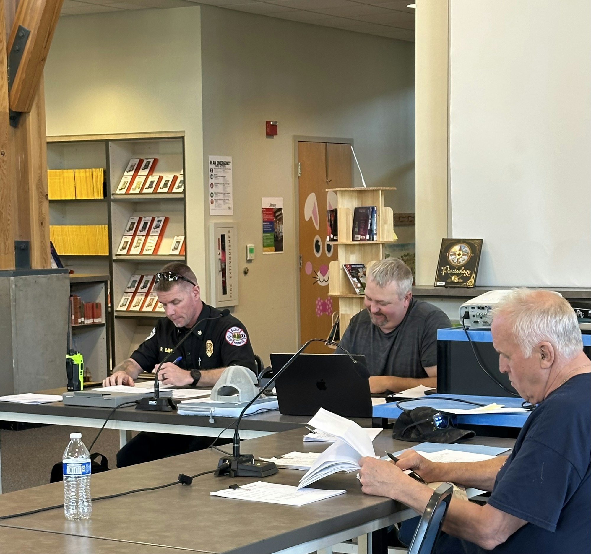 Three men sitting at tables with papers and laptops, in a library or office setting. Shelves and a decorated door are in the background.
