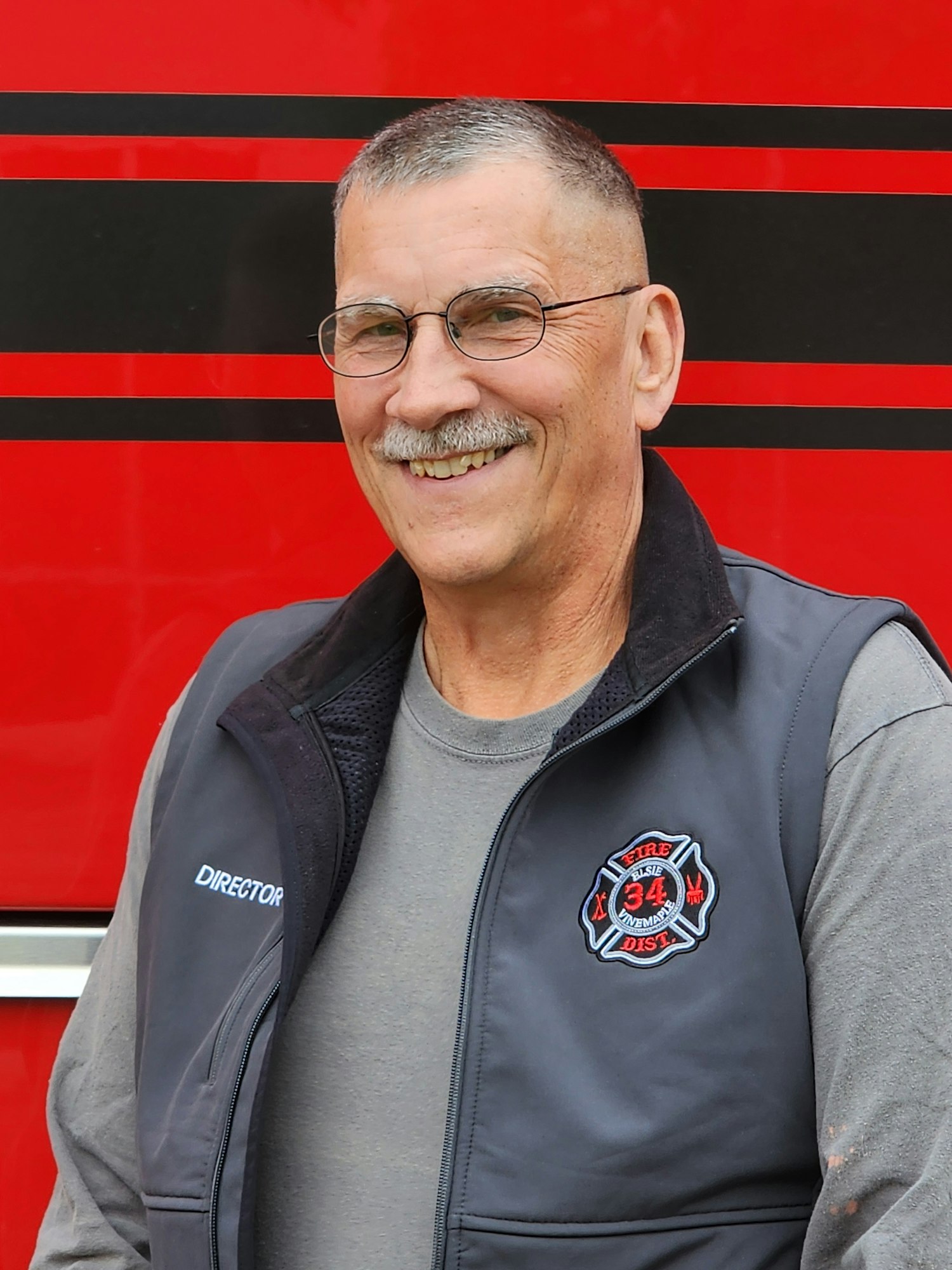 A person smiling, wearing glasses and a vest labeled "Director" with a fire department logo, in front of a red background.
