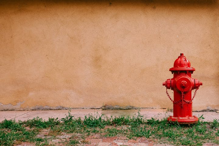 A red fire hydrant against a beige wall with some grass and a brick surface at the bottom.