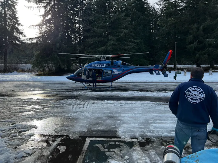 A helicopter is preparing for takeoff in a snowy area, with a person in a blue jacket observing nearby.