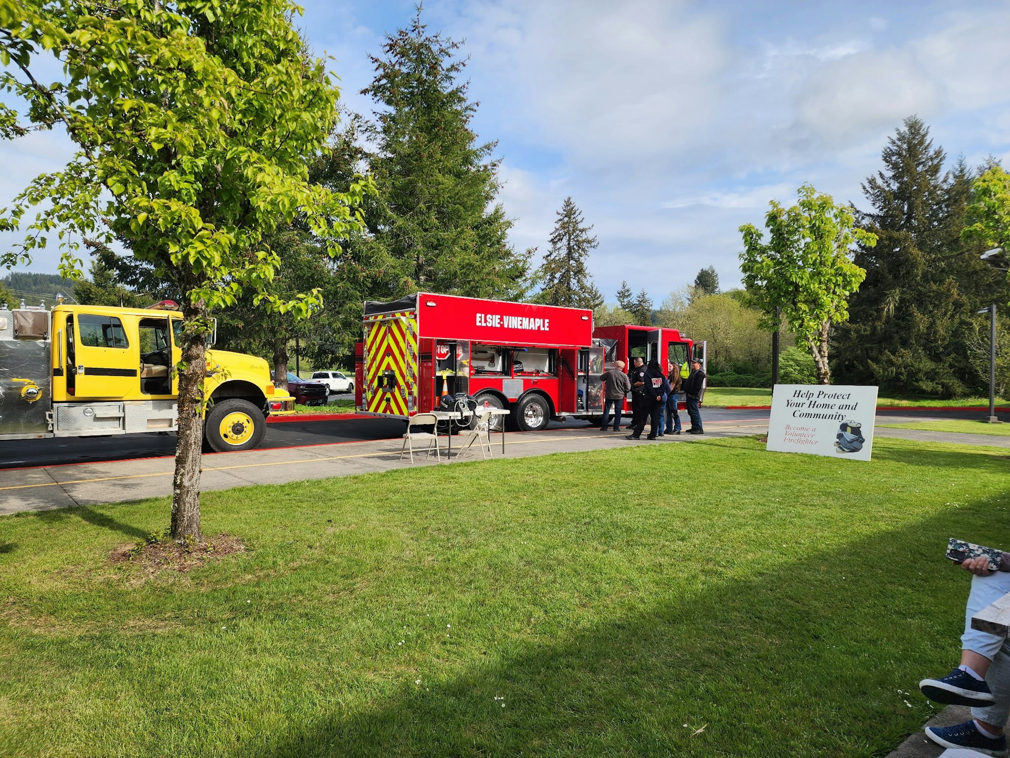 Firetruck and crew outdoors with a "help protect" sign, chairs, and trees.