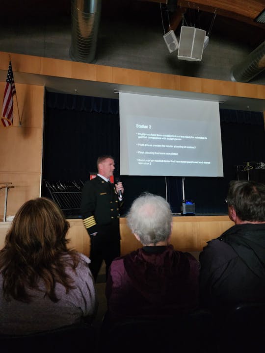 A person in a uniform presents in front of an audience, with a projected slide titled "Station 2" in a dimly lit room.