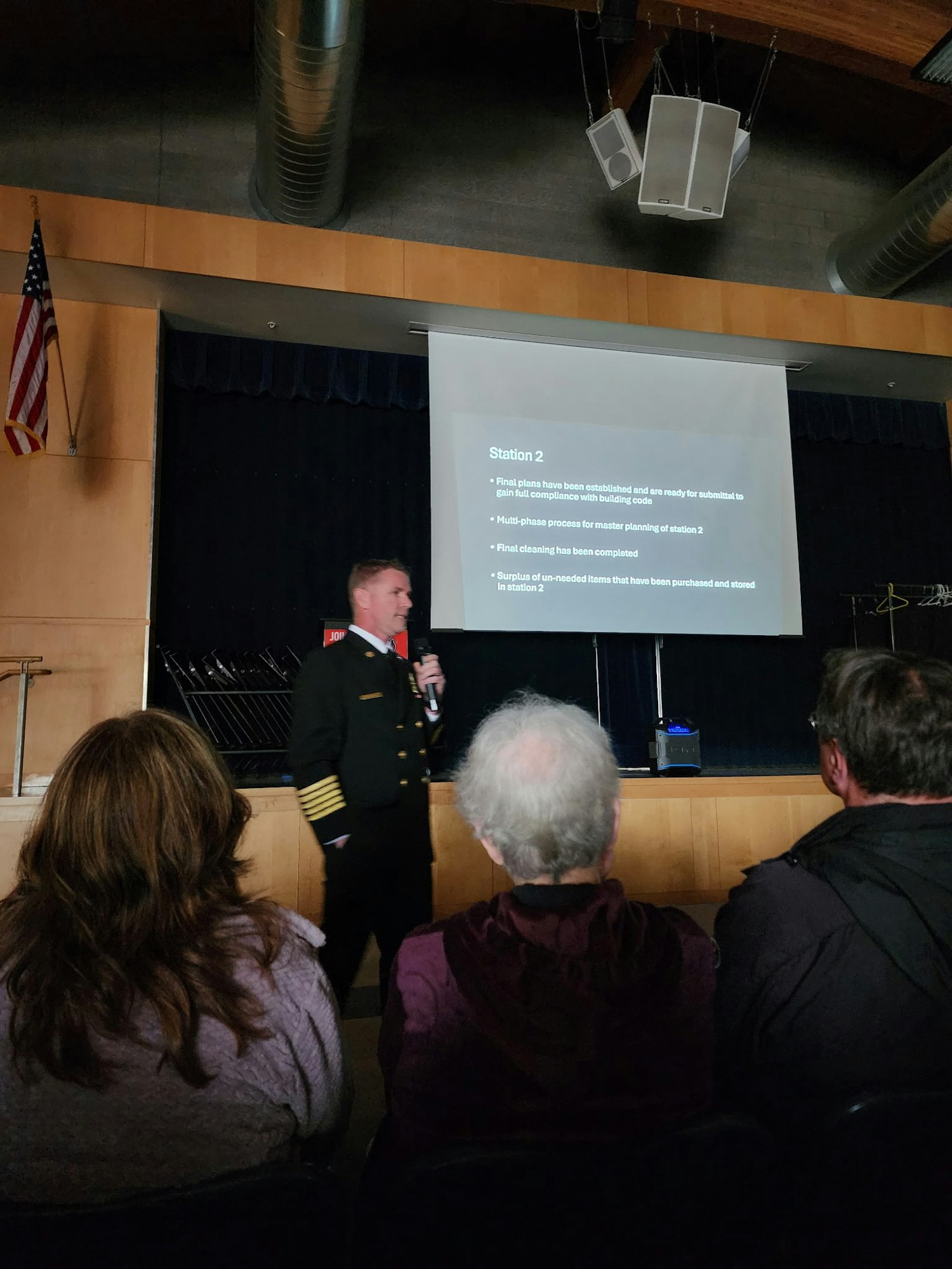 A person in a uniform presents in front of an audience, with a projected slide titled "Station 2" in a dimly lit room.