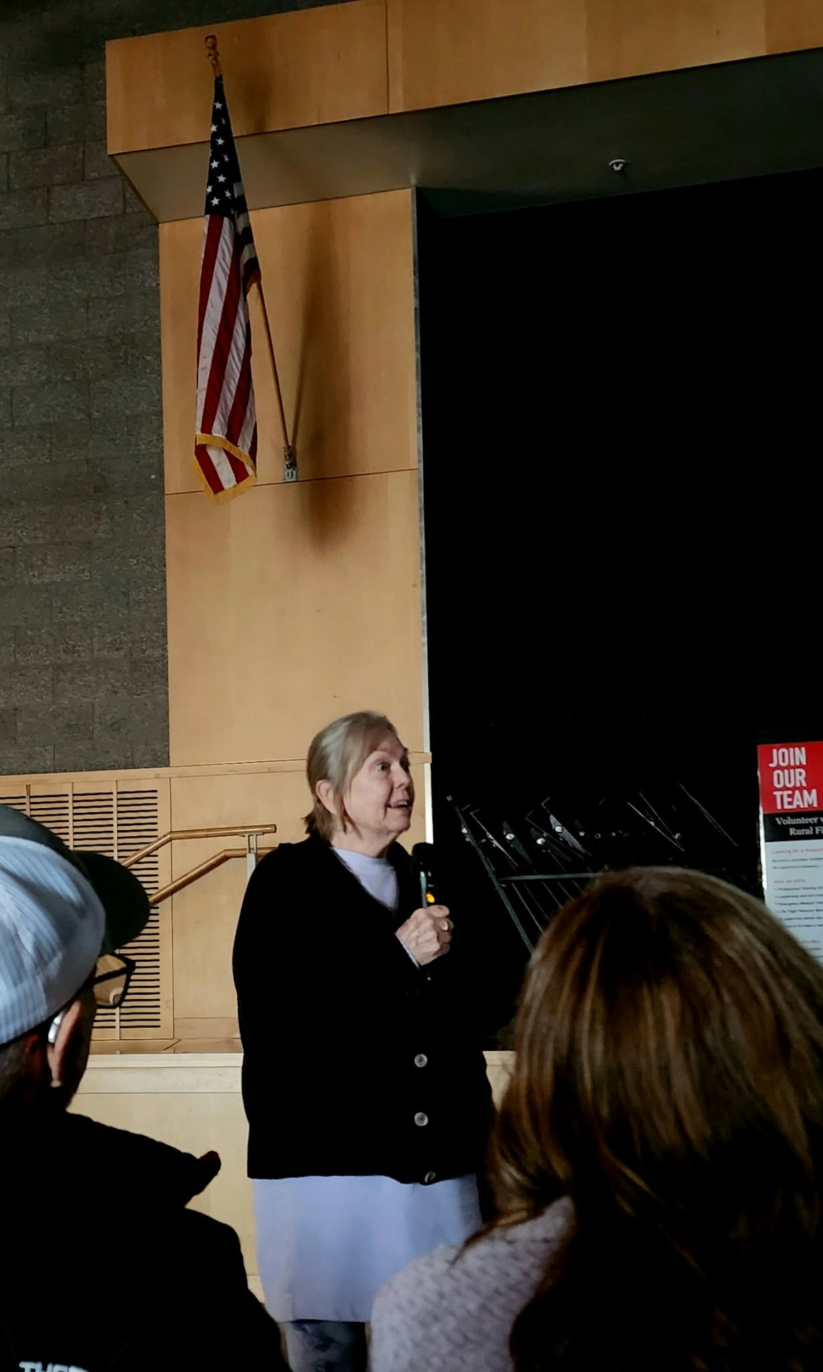 A person speaking on stage with a microphone, an American flag on the wall, and "Join Our Team" text on a poster.