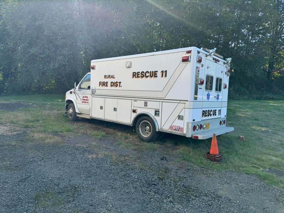 Ambulance labeled "RESCUE 11" from a rural fire district parked on a gravel area with trees and a traffic cone beside it.