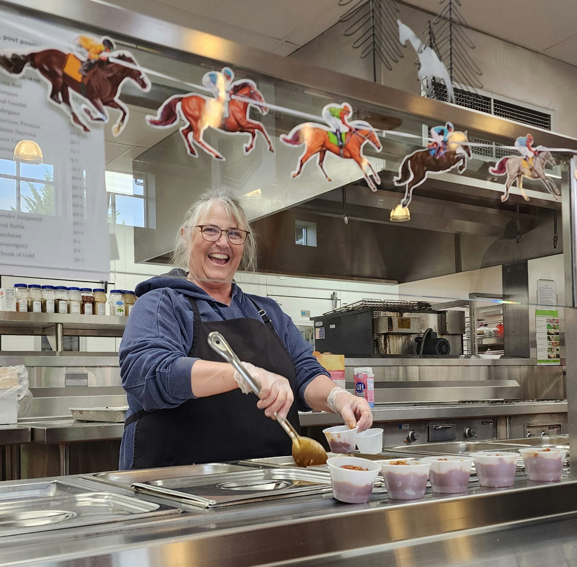 A smiling person serving food in a kitchen with horse decorations above.