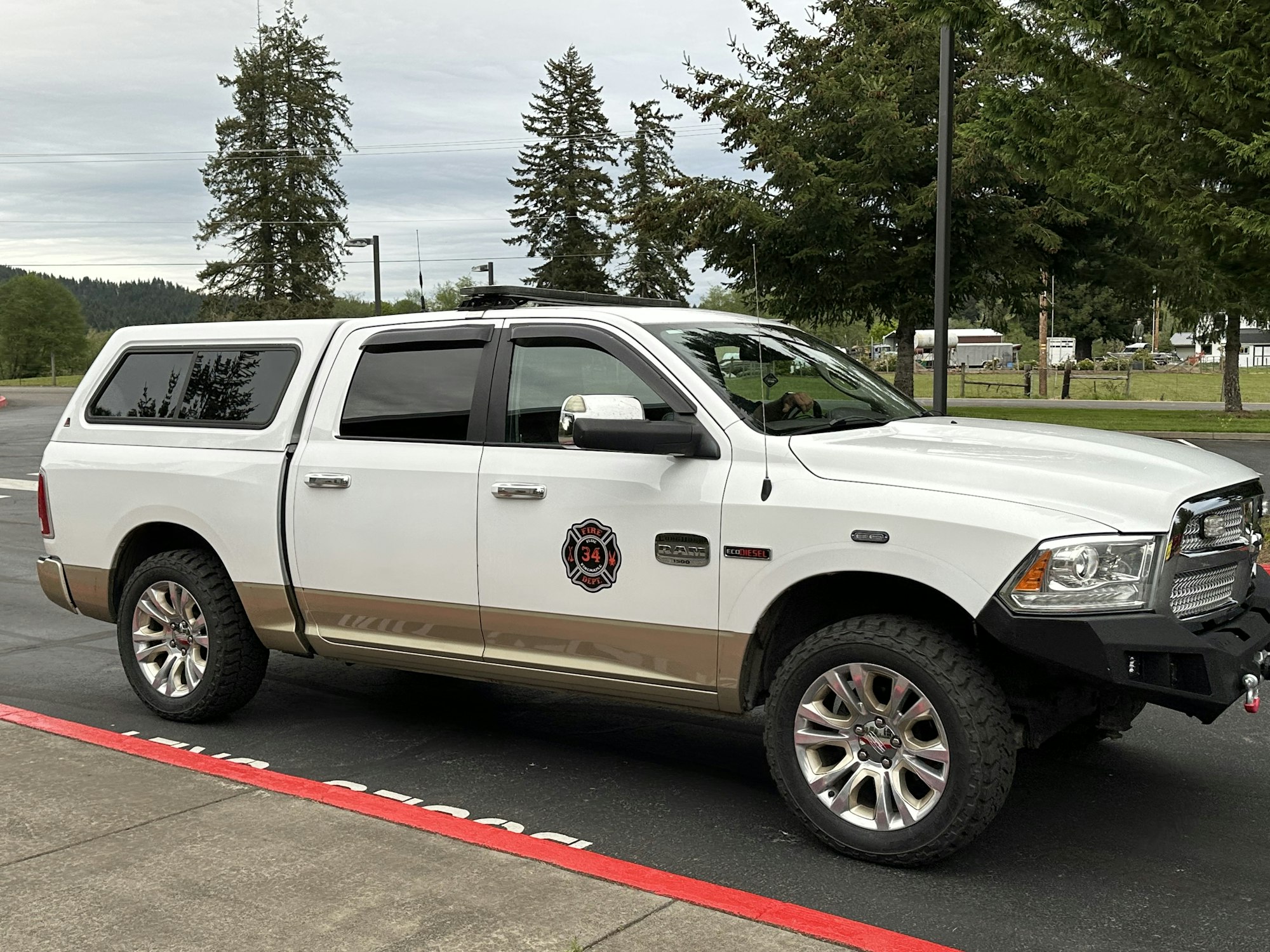 A white pickup truck with a fire department emblem is parked on a paved area next to a red line, surrounded by trees.