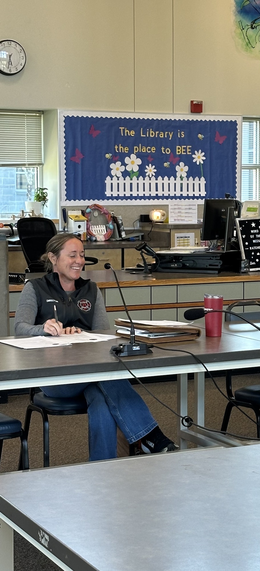 A person smiling at a table with papers and a microphone, in a room with a library-themed bulletin board.