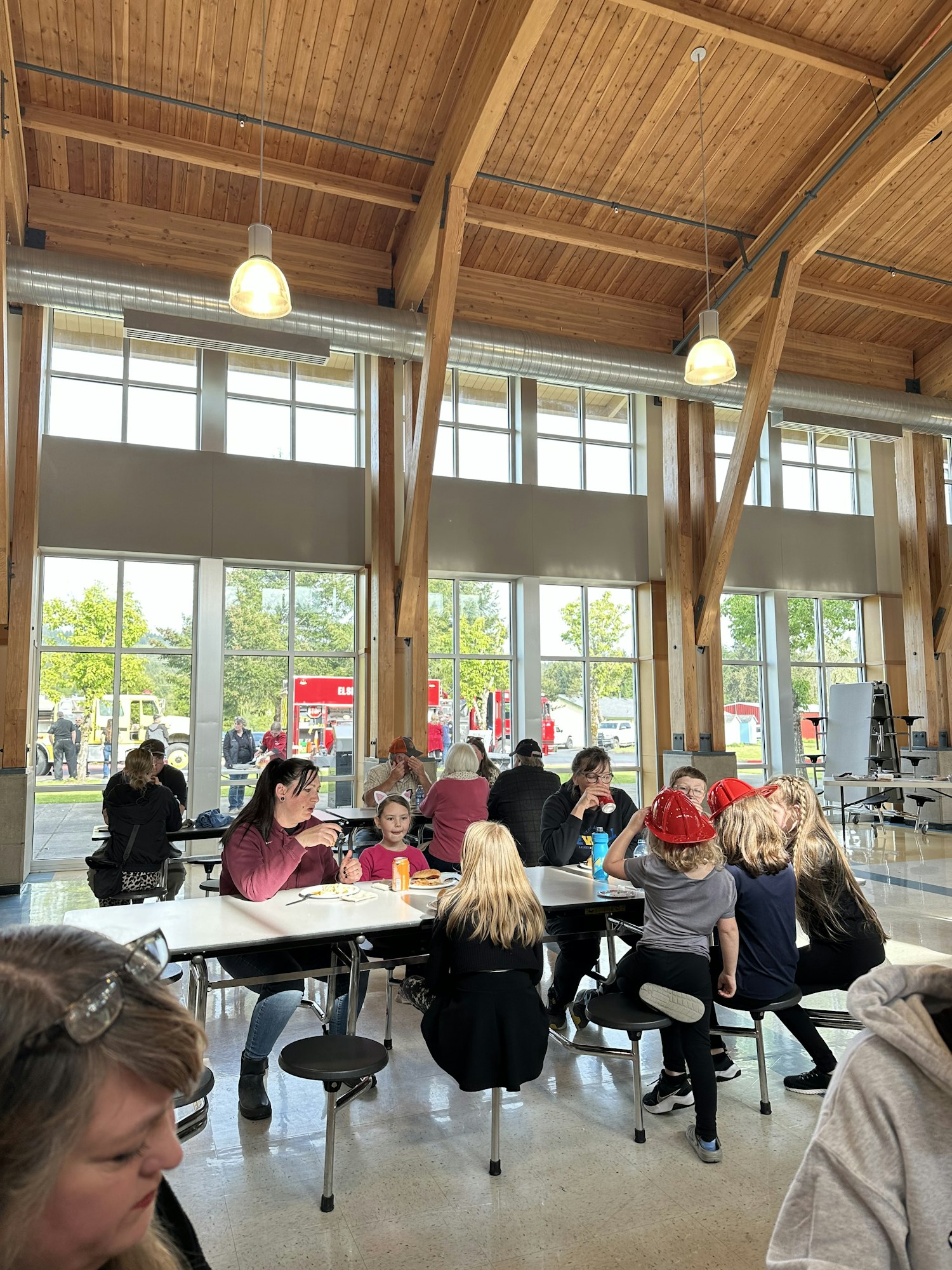 A group of people sitting at tables in a bright, wooden ceiling room with large windows, some wearing red hats.