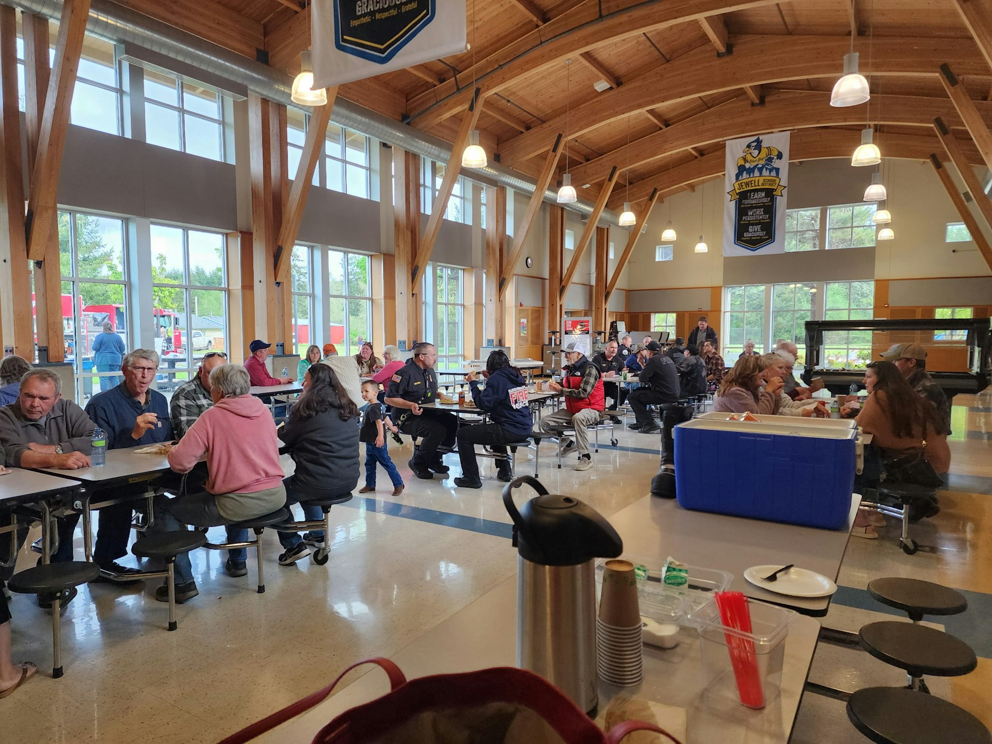 People sitting and eating at tables in a large hall with high ceilings and windows. A banner and blue cooler are visible.