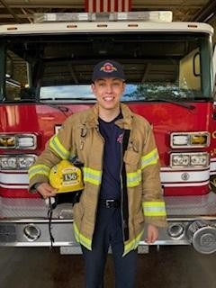 A smiling person in firefighter gear holding a helmet, standing in front of a fire truck.