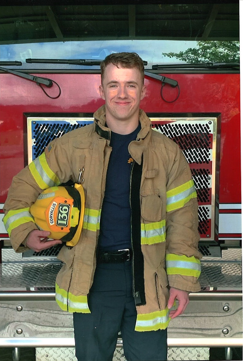A smiling person in firefighter gear holding a helmet, standing in front of a fire truck.