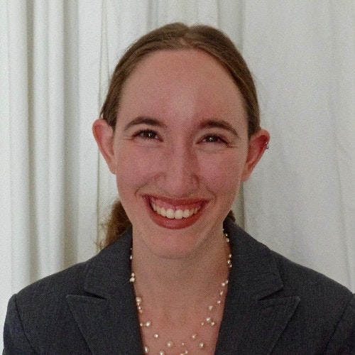 A person smiling, wearing a blazer and pearl necklace, against a white background.