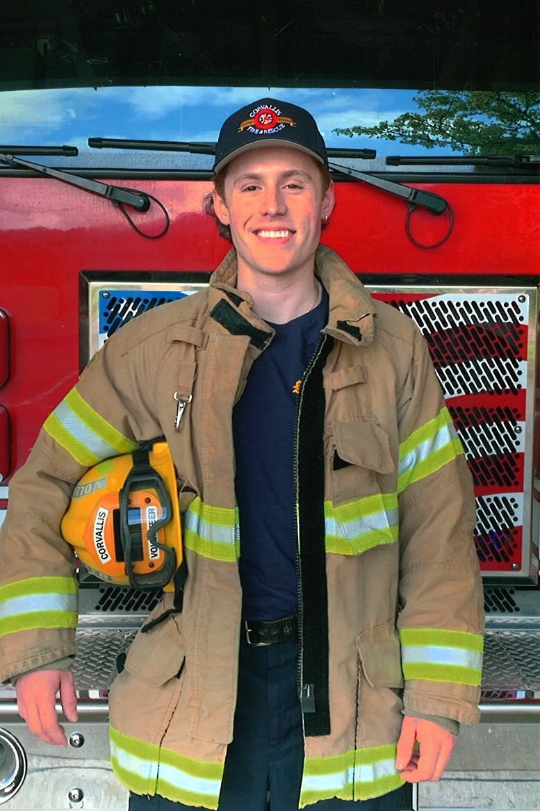 A smiling firefighter in gear standing in front of a fire truck.