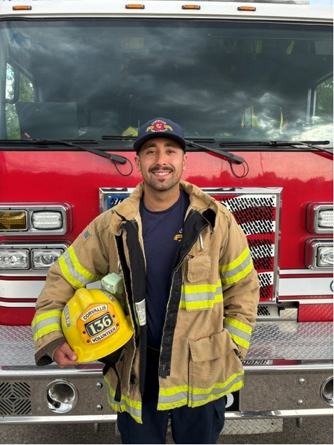A smiling firefighter in a protective uniform stands in front of a fire truck, holding a yellow helmet with the number 136.
