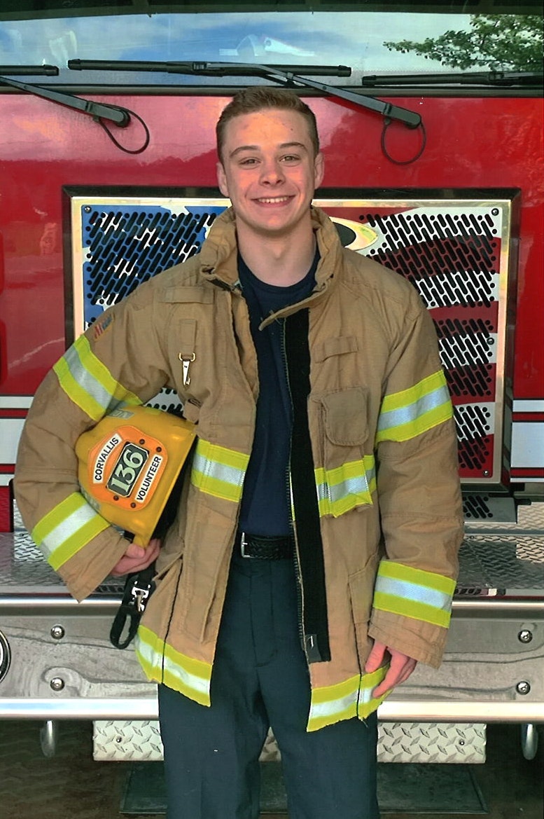 A smiling person in firefighter turnout gear holding a helmet, standing in front of a fire truck.