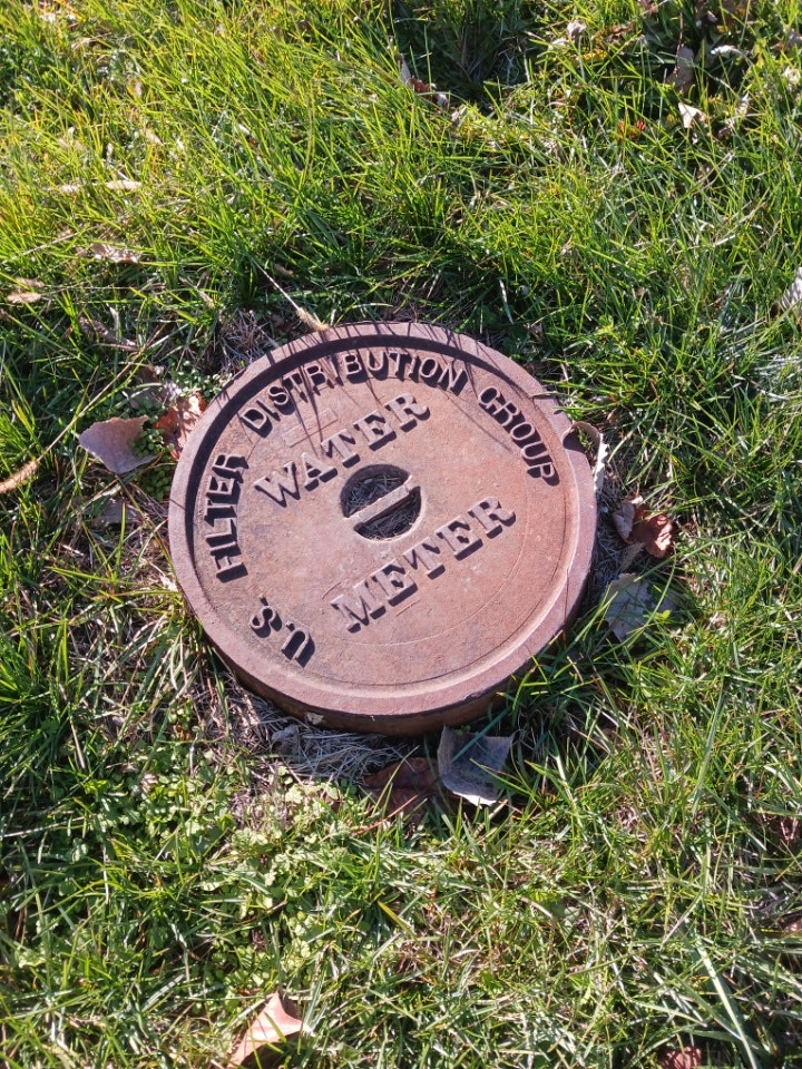 A round metal water meter cover labeled "SS & Filter Distribution Group" in grass.