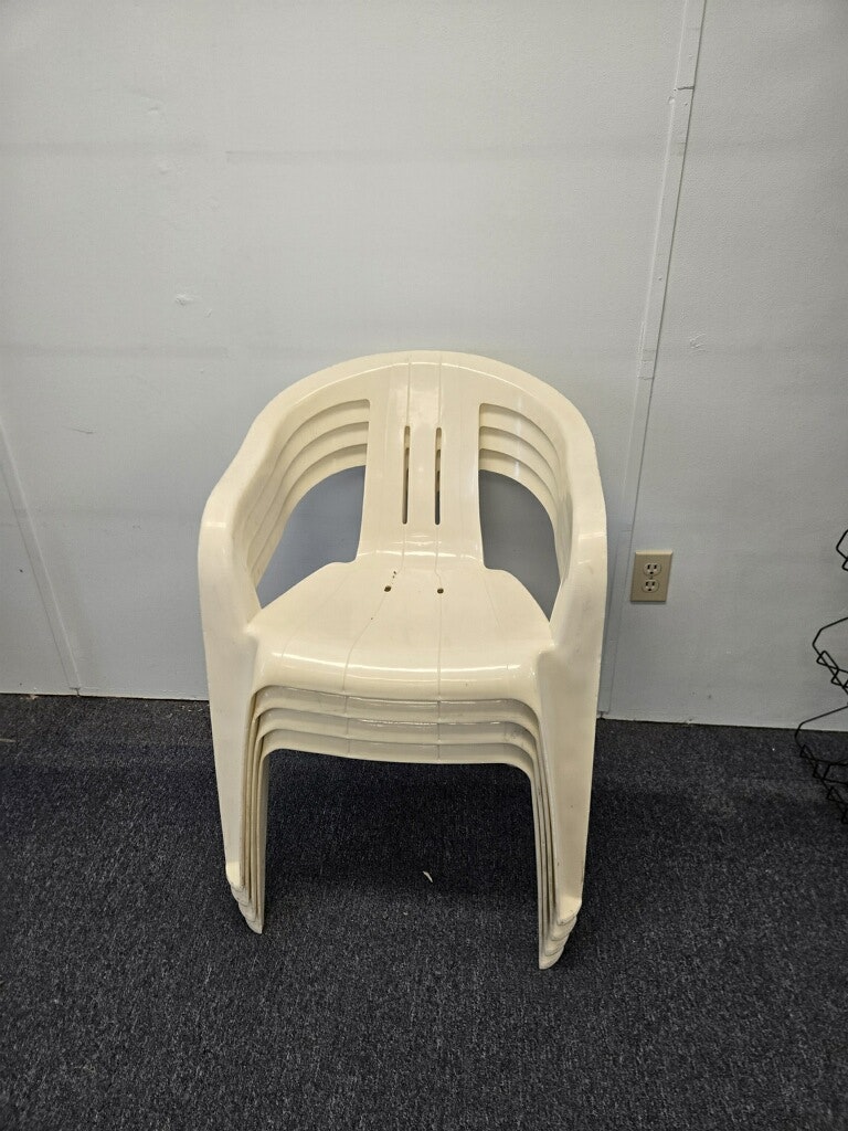 A stack of four white plastic chairs arranged neatly on a dark carpeted floor, against a plain wall.