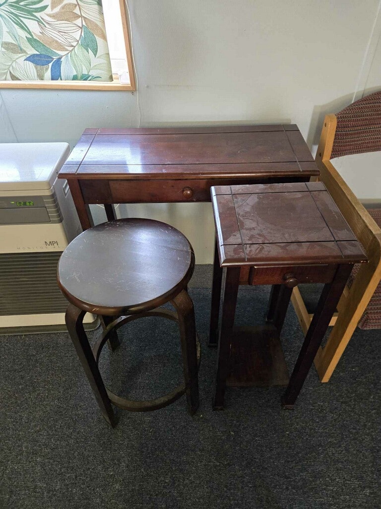 A small wooden table, a stool, and a nightstand are arranged together in a room with a window and a corner chair.