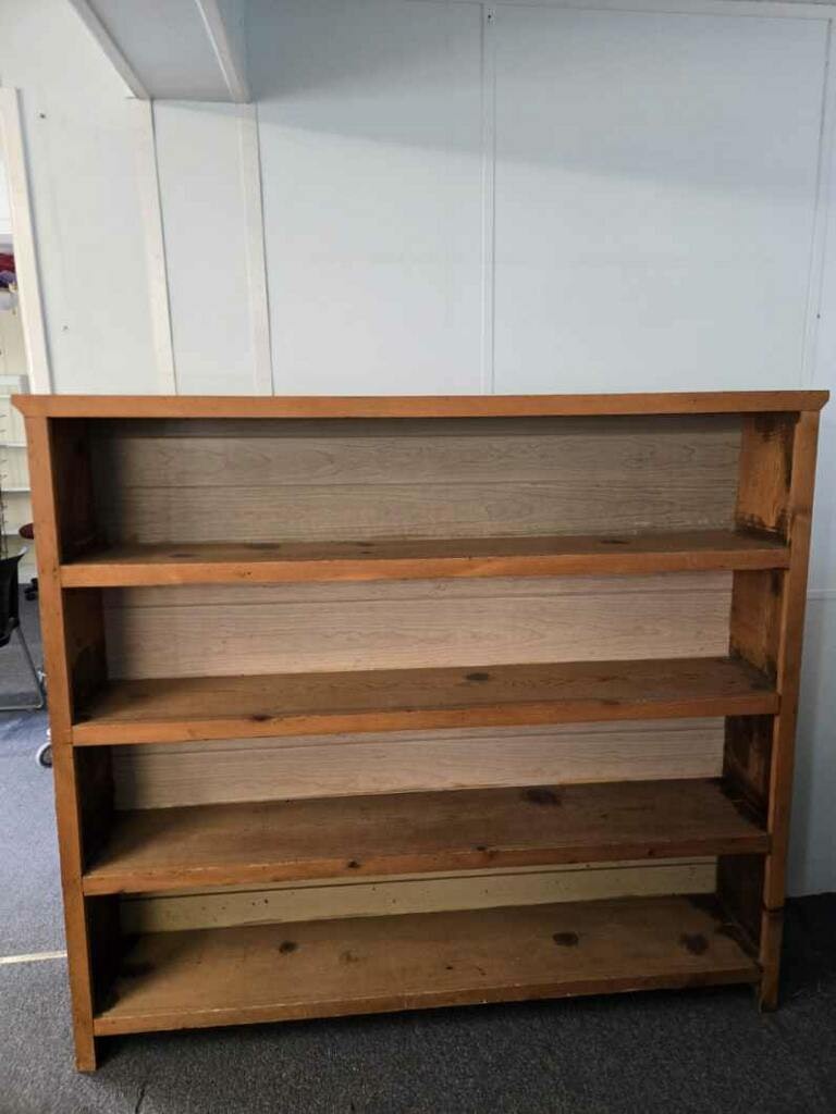The image shows a wooden bookshelf with four shelves, placed against a wall in a room.