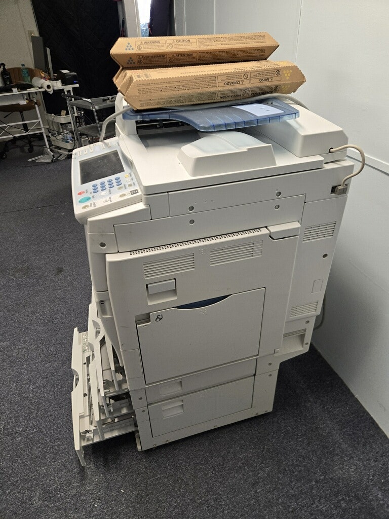 A multifunction printer/copier with two cardboard boxes on top, set in a room with dark carpet and dim lighting.