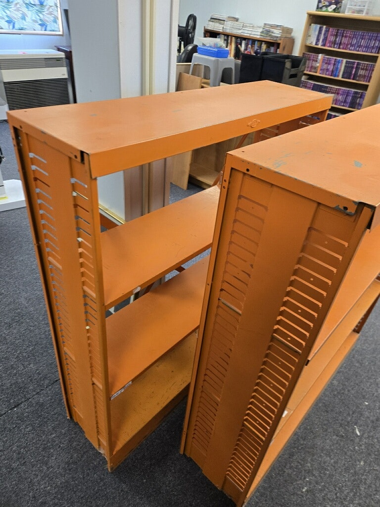 The image shows two orange metal shelves, standing indoors, with a background of bookshelves and carpeted floor.