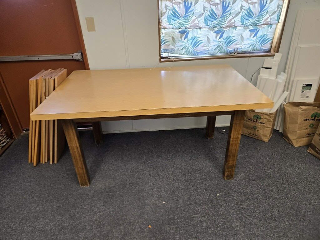 A simple wooden table with a light-colored top and four wooden legs, located in a room with carpet and window curtains.