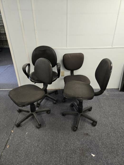 The image shows four black office chairs with wheels, varying in style and height, placed against a light-colored wall.