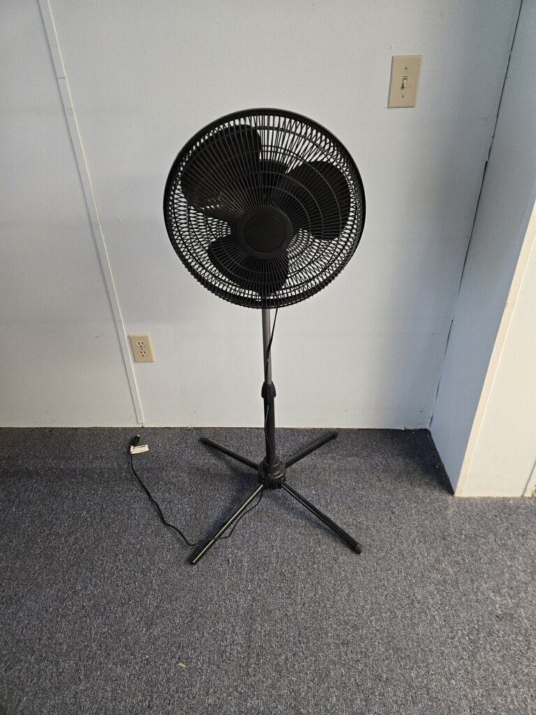 A black standing fan positioned on a flat surface, with a power cord visible nearby, in a simple indoor setting.