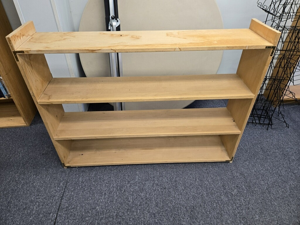 A simple, unfinished wooden bookshelf with four shelves, located on a carpeted floor.
