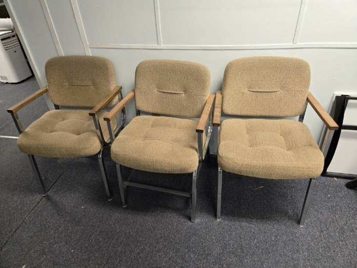Three beige fabric chairs with metal frames and wooden armrests are aligned against a wall on a carpeted floor.