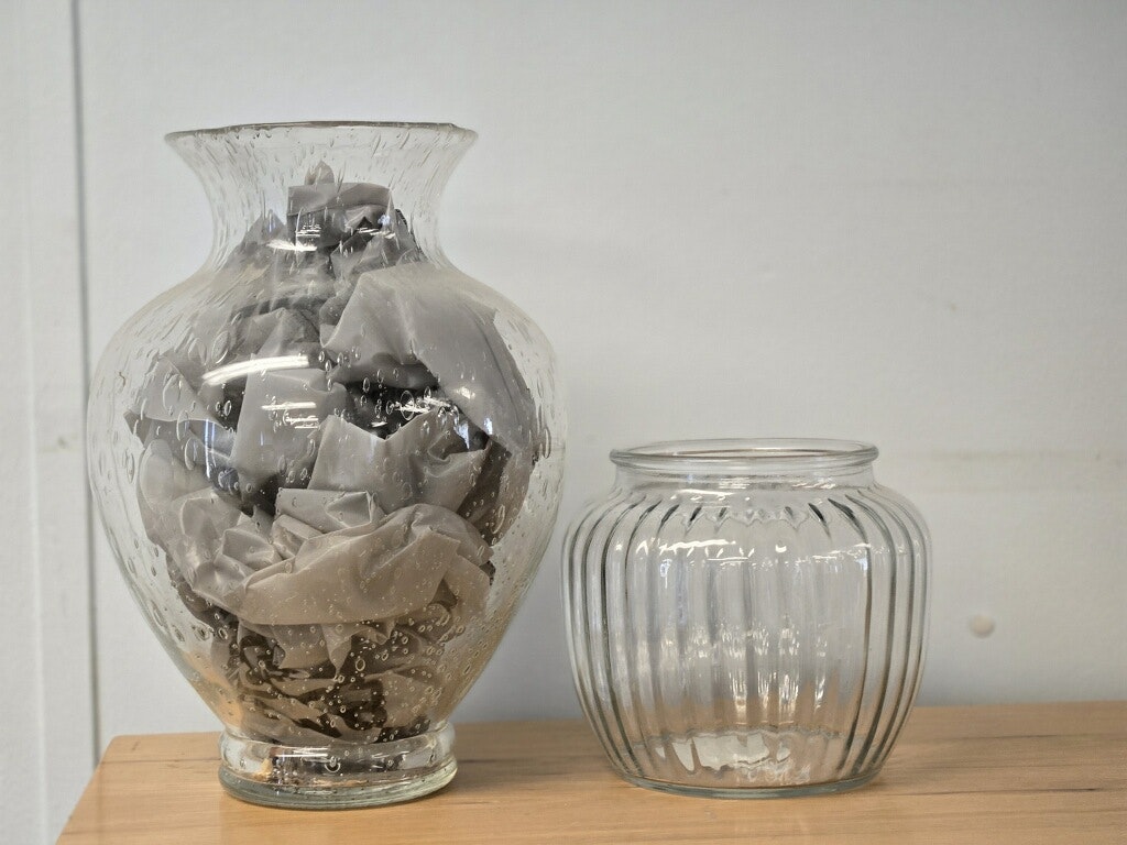 The image showcases two glass containers: a larger vase filled with folded gray fabric and a smaller ribbed glass jar.