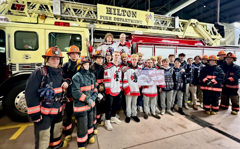 A group of people in hockey jerseys and firefighter gear stand in front of a Hilton Fire Department truck, some holding a sign.