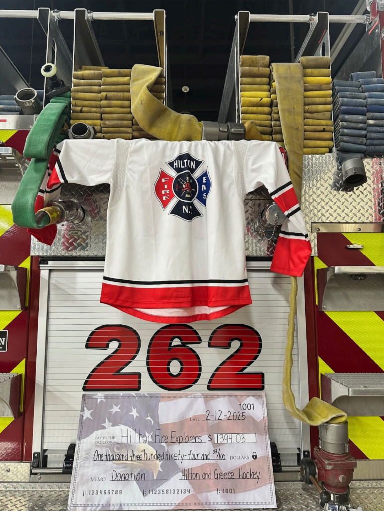 Fire gear with a sports jersey draped on it. A large check for $1,394.03 is addressed to Hilton Fire Explorers from Hilton and Greece Hockey.