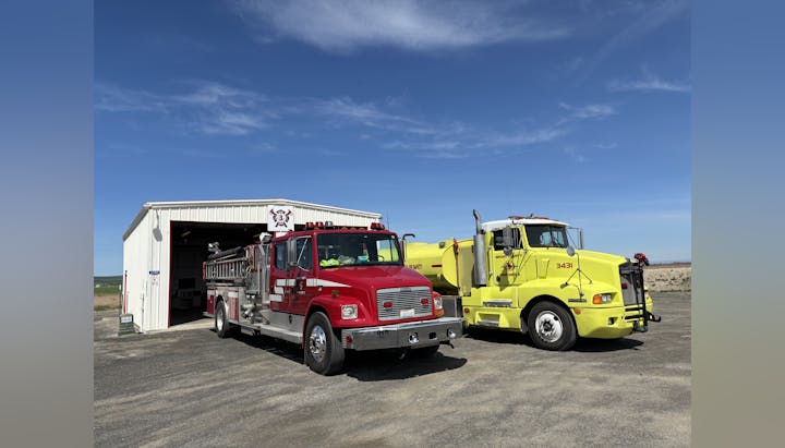 Two fire trucks parked outside a small fire station under a clear blue sky.