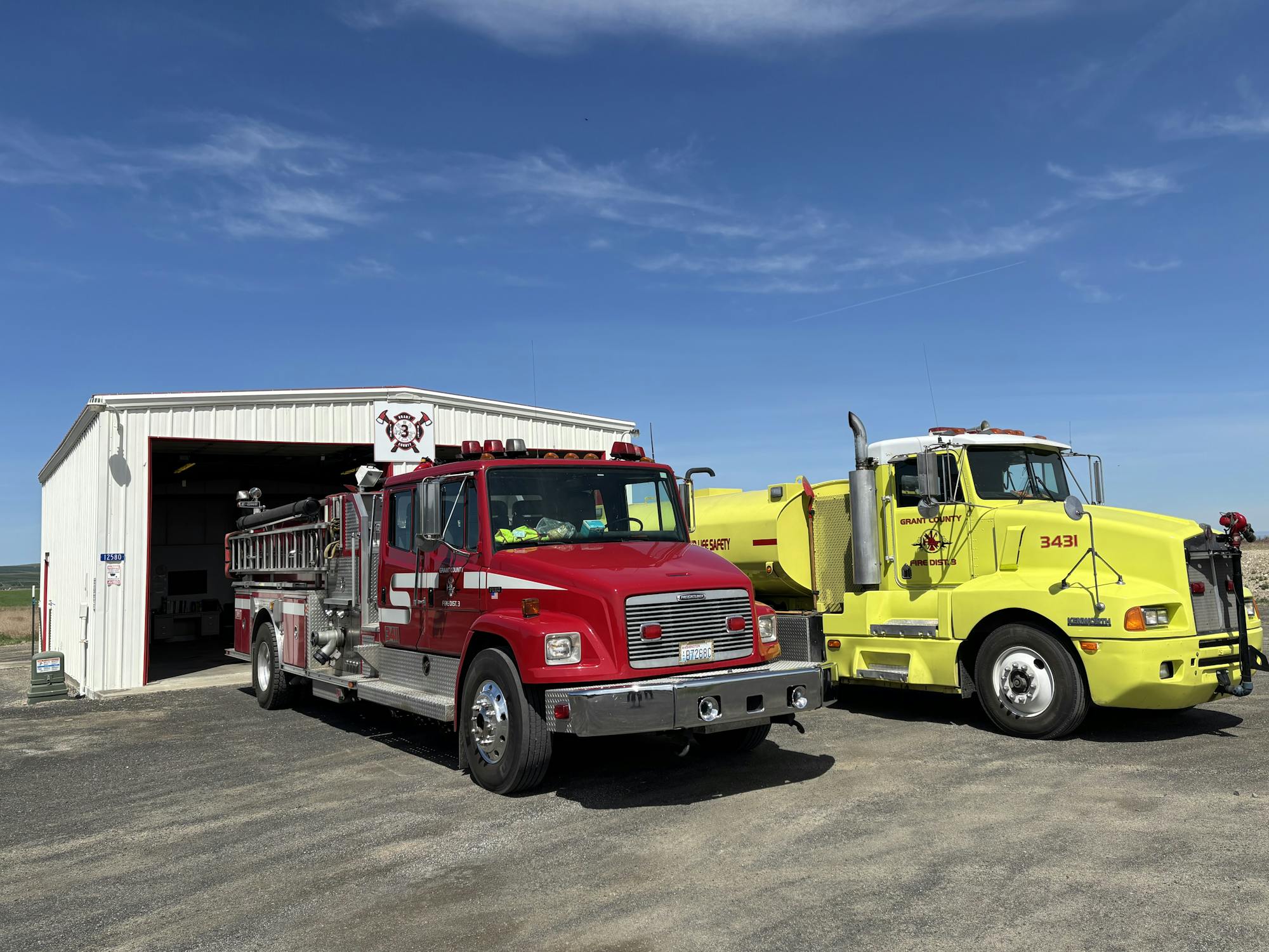Two fire trucks parked outside a small fire station under a clear blue sky.