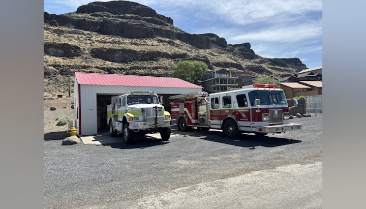 Two fire trucks outside a small fire station with a red roof, set against a rocky hillside.