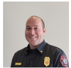 A person in a firefighter uniform smiles at the camera, with a badge and embroidered emblem visible on the shirt.