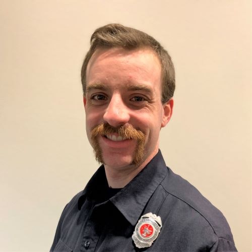 A person with a mustache in a uniform, wearing a badge, smiling.