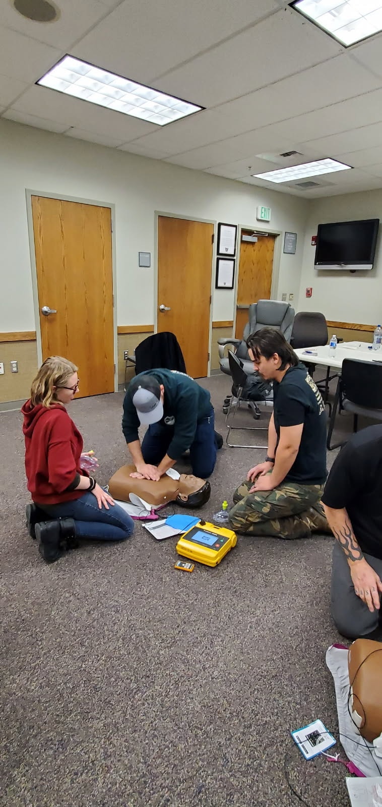 A CPR training session with three people practicing on a manikin, using an AED in a classroom setting.