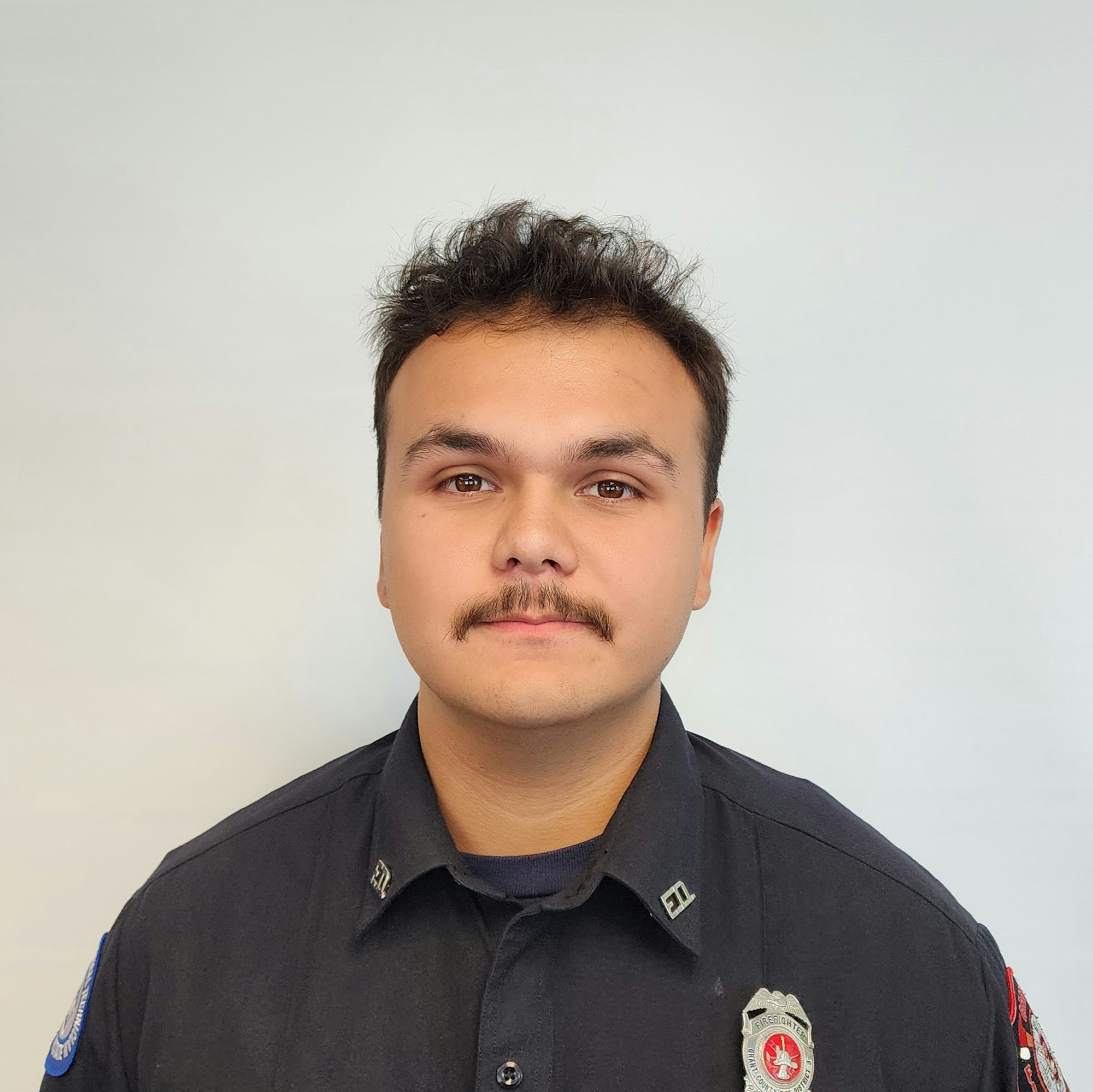 A person with a mustache wearing a dark uniform and badge, looking directly at the camera against a plain background.