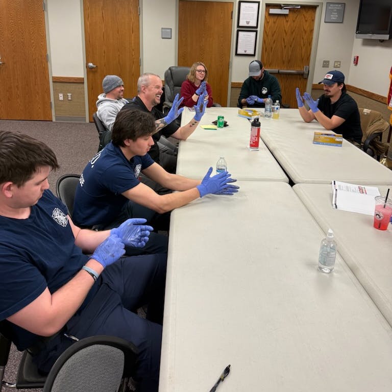 A group of people sit around a table wearing blue gloves, engaging in discussion.